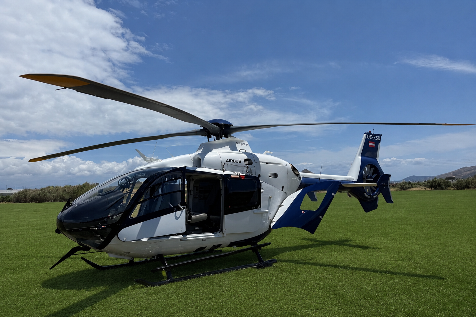 Airbus H135 helicopter parked at Paros island helipad with green landing ground under blue Aegean sky